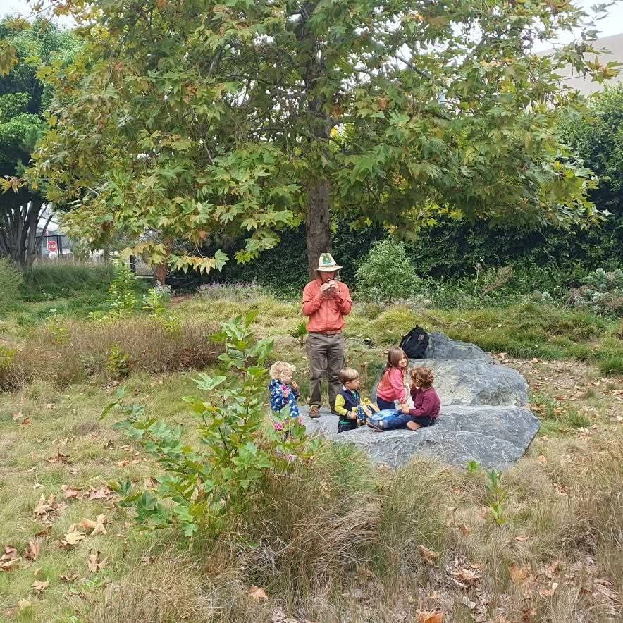 Philip and four young children pausing on a large rock beneath a sycamore, in a grassy patch at the edge of the city